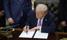 Trump sits at his desk in the Oval Office. He is leaned over, signing an executive order. 