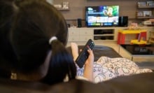 A young girl watches TV with remote in her hand.
