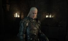 A man in armour stands in a dark room lit by two burning torches.