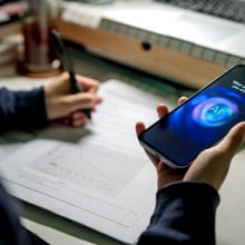 A teen holds a phone in their hand and consults an AI for help while writing in a notebook. 