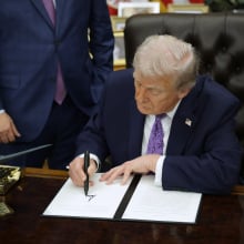 Trump sits at his desk in the Oval Office. He is leaned over, signing an executive order. 