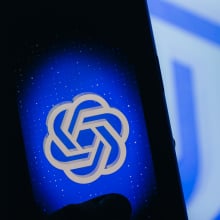 A hand in shadows holds a glowing phone, displaying a blue ChatGPT logo.
