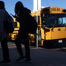 Students holding cellphones walk by a yellow school bus.