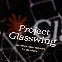 The visual for Project Glasswing is displayed on a smartphone screen placed on a reflective surface onto which a Claude logo with hexagonal patterns is projected