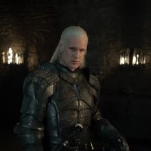 A man in armour stands in a dark room lit by two burning torches.