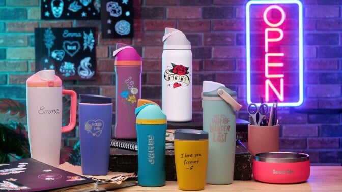the Owala Tattoos customization options on many water bottles and a dog bowl, displayed on a table together with a neon "open" sign in the background