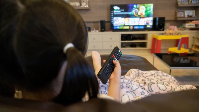 A young girl watches TV with remote in her hand.