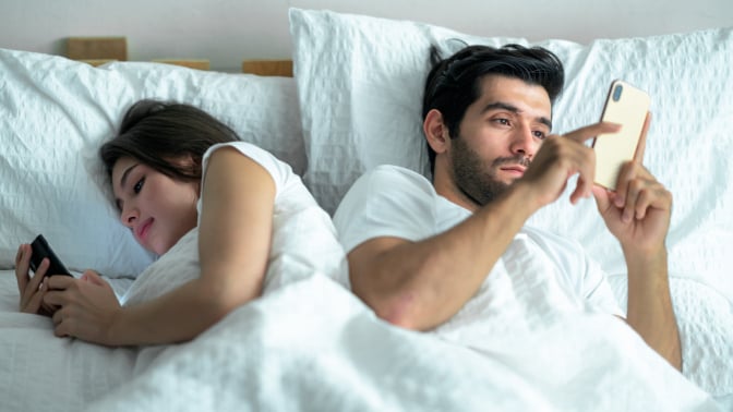 young couple using on their phones in bed back to back