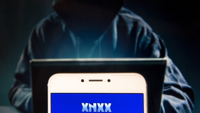 In this photo illustration, the Pornographic video sharing and viewing websites Xnxx logo is seen displayed on an Android mobile device with a figure of hacker in the background