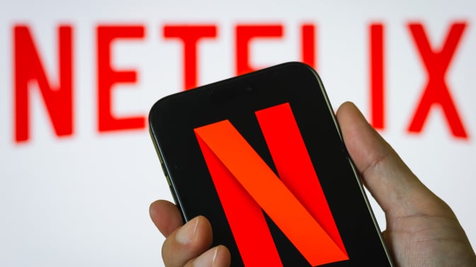  In this photo illustration, a person holds a smartphone in vertical orientation displaying the red N logo of Netflix, Inc. (NASDAQ: NFLX) in front of a blurred background showing the company name in bold red letters on July 13, 2025.