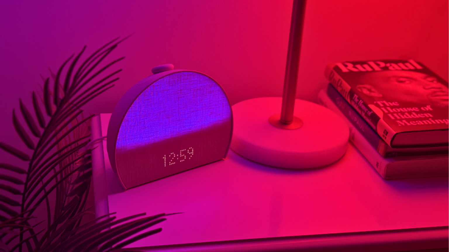 Hatch Restore 3 alarm clock glowing purple showing 12:59 on clock, sitting on nightstand near plant and lamp