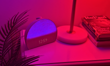 Hatch Restore 3 alarm clock glowing purple showing 12:59 on clock, sitting on nightstand near plant and lamp