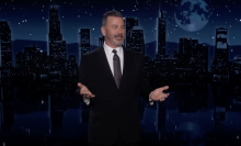 A man in a suit stands on a talk show stage, arms wide and grinning.
