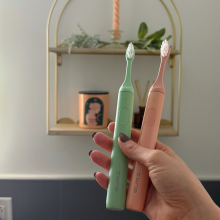 Hand holding pink and green Suri toothbrushes in front of bathroom wall