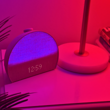 Hatch Restore 3 alarm clock glowing purple showing 12:59 on clock, sitting on nightstand near plant and lamp