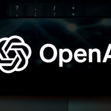 The OpenAI logo appears on the screen of a smartphone placed on a laptop keyboard illuminated by green light.