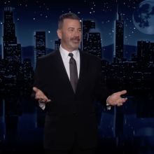 A man in a suit stands on a talk show stage, arms wide and grinning.