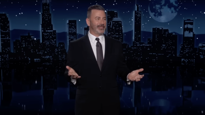 A man in a suit stands on a talk show stage, arms wide and grinning.